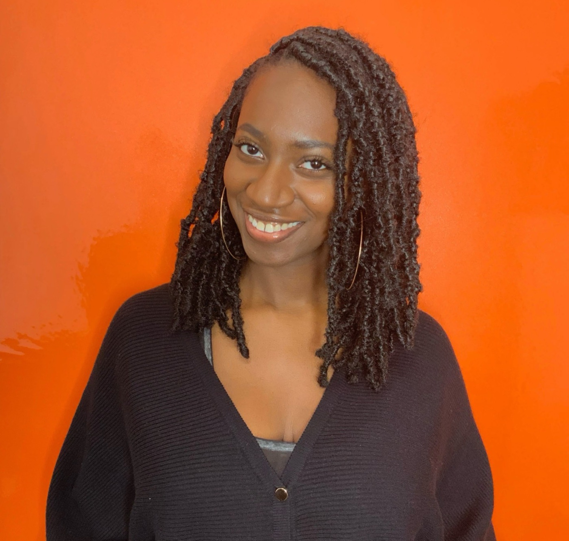 A smiling woman with shoulder-length locs and gold hoop earrings, photographed against a bright orange background.