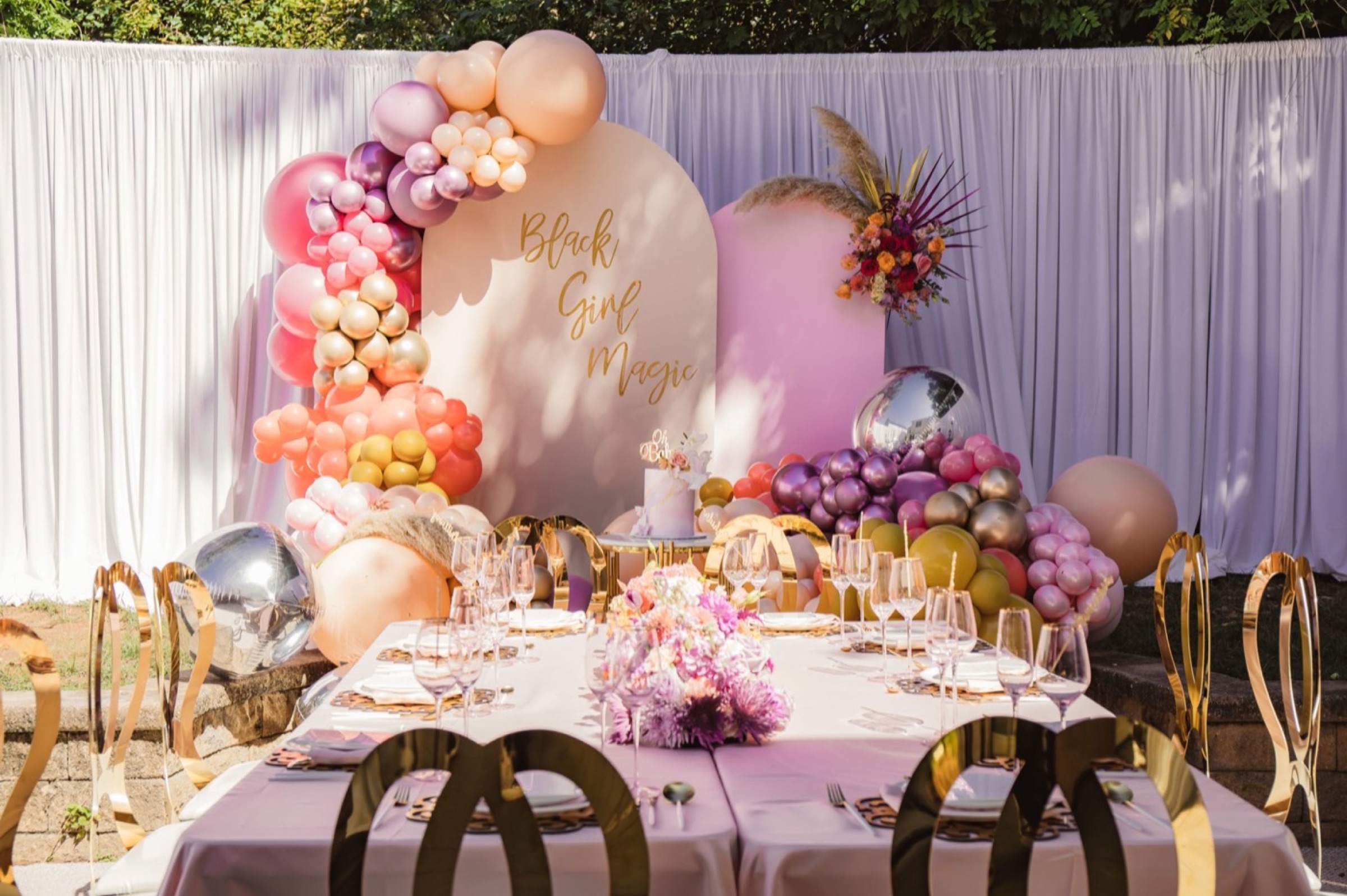 A baby shower backdrop of pink, peach and mauve balloons around an arch reading Black Girl Magic, with a set table in front.