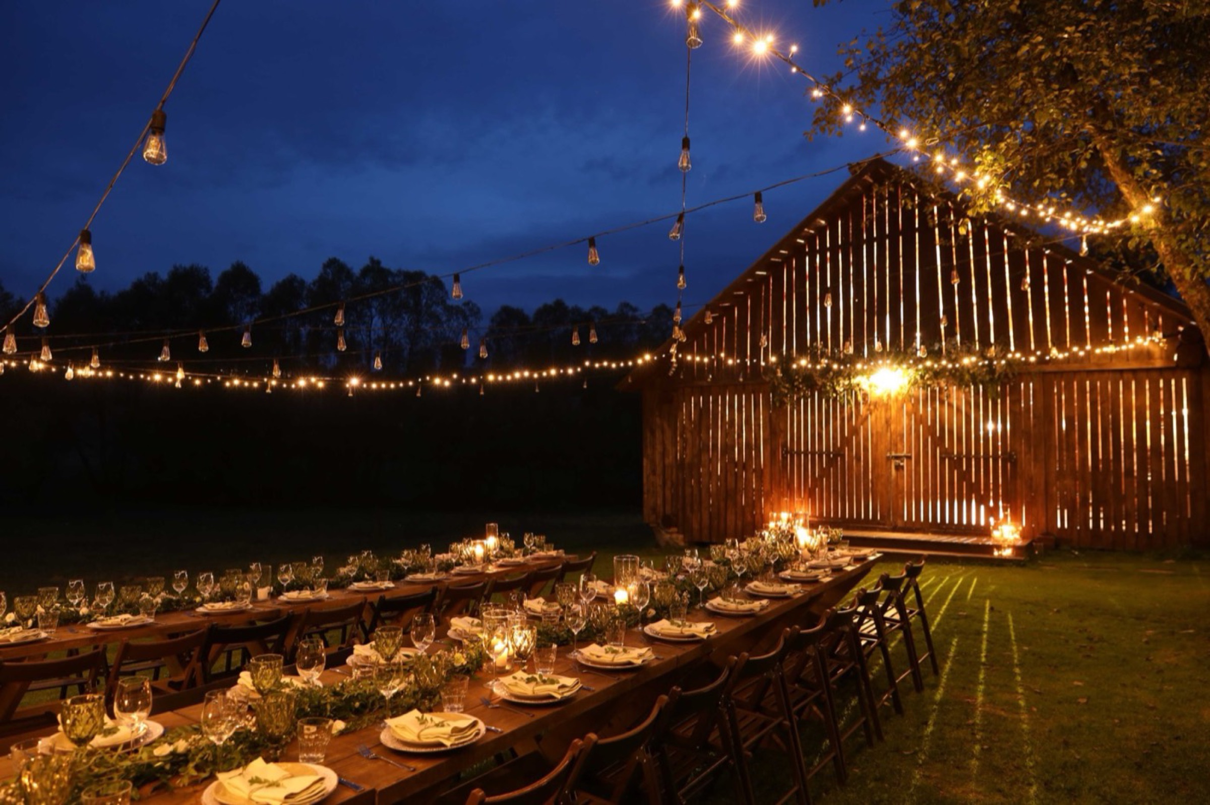 A long banquet table set for dinner outside a timber barn at dusk, lit by strings of bulbs overhead.
