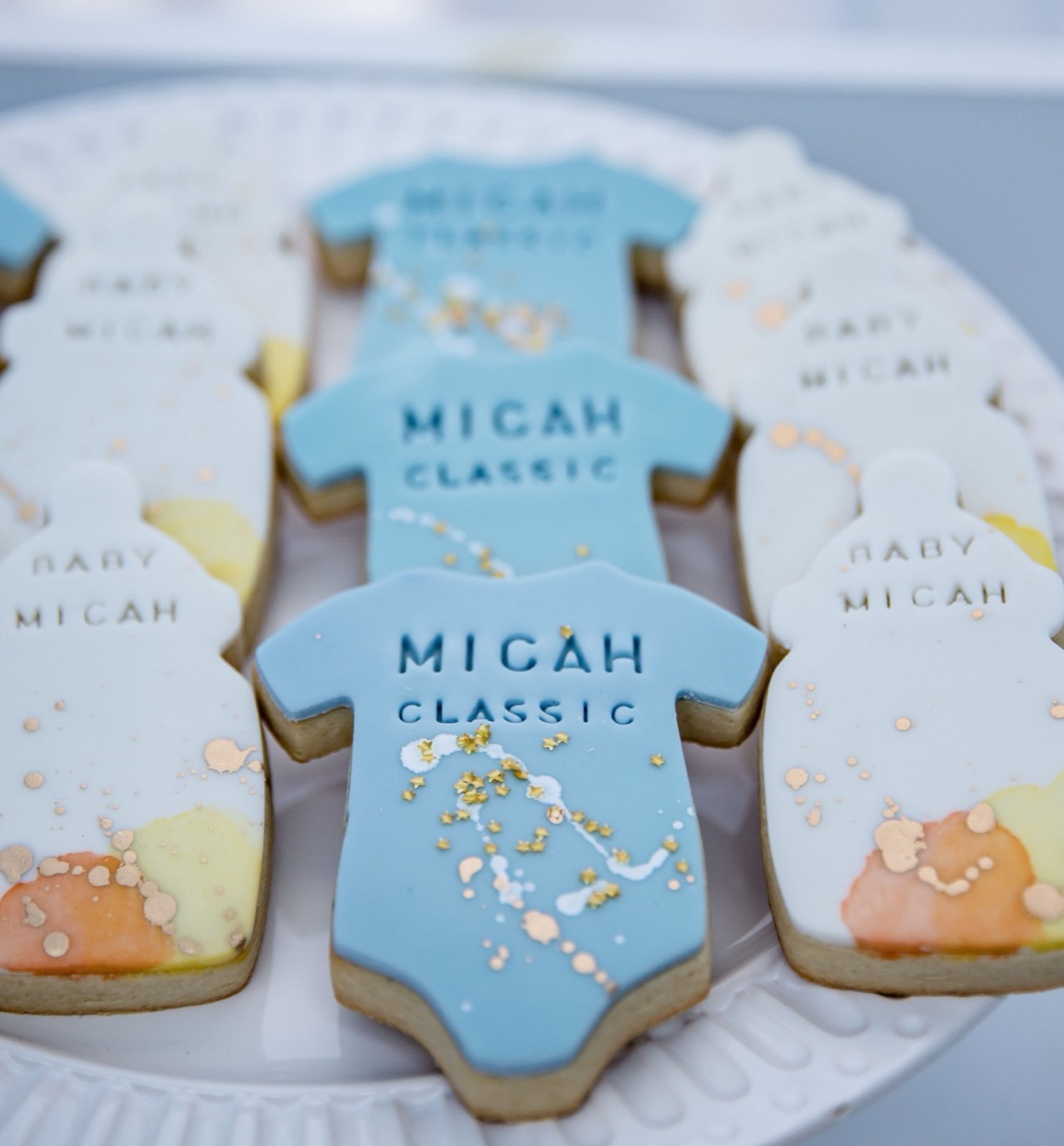 Iced sugar cookies shaped like baby onesies, decorated in pale blue and gold and lettered Micah Classic and Baby Micah.