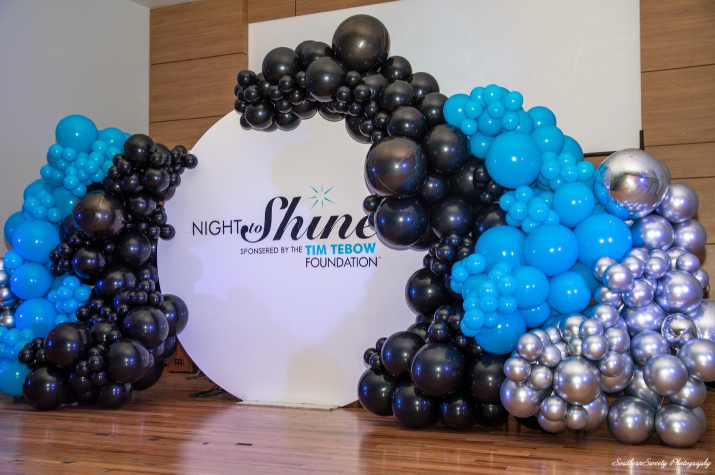 A circular white backdrop framed by an arch of black, blue and silver balloons, printed Night to Shine, sponsored by the Tim Tebow Foundation.
