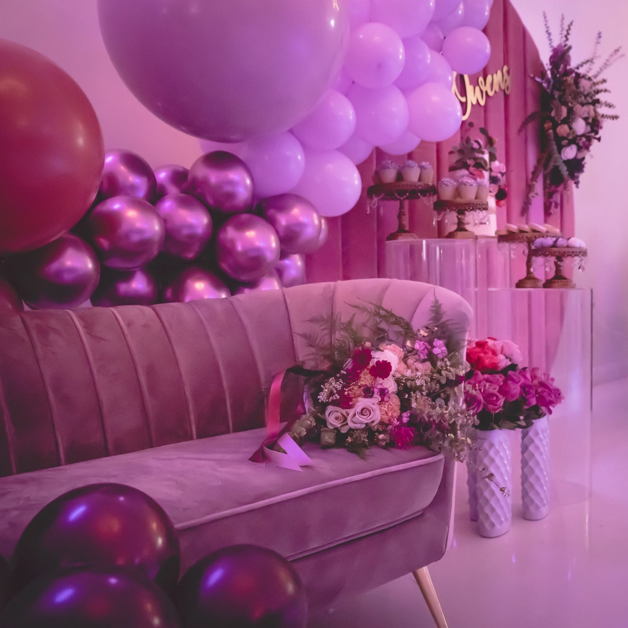 A baby shower lounge set in shades of purple: a velvet sofa banked with balloons, a floral arrangement and a tiered dessert display.