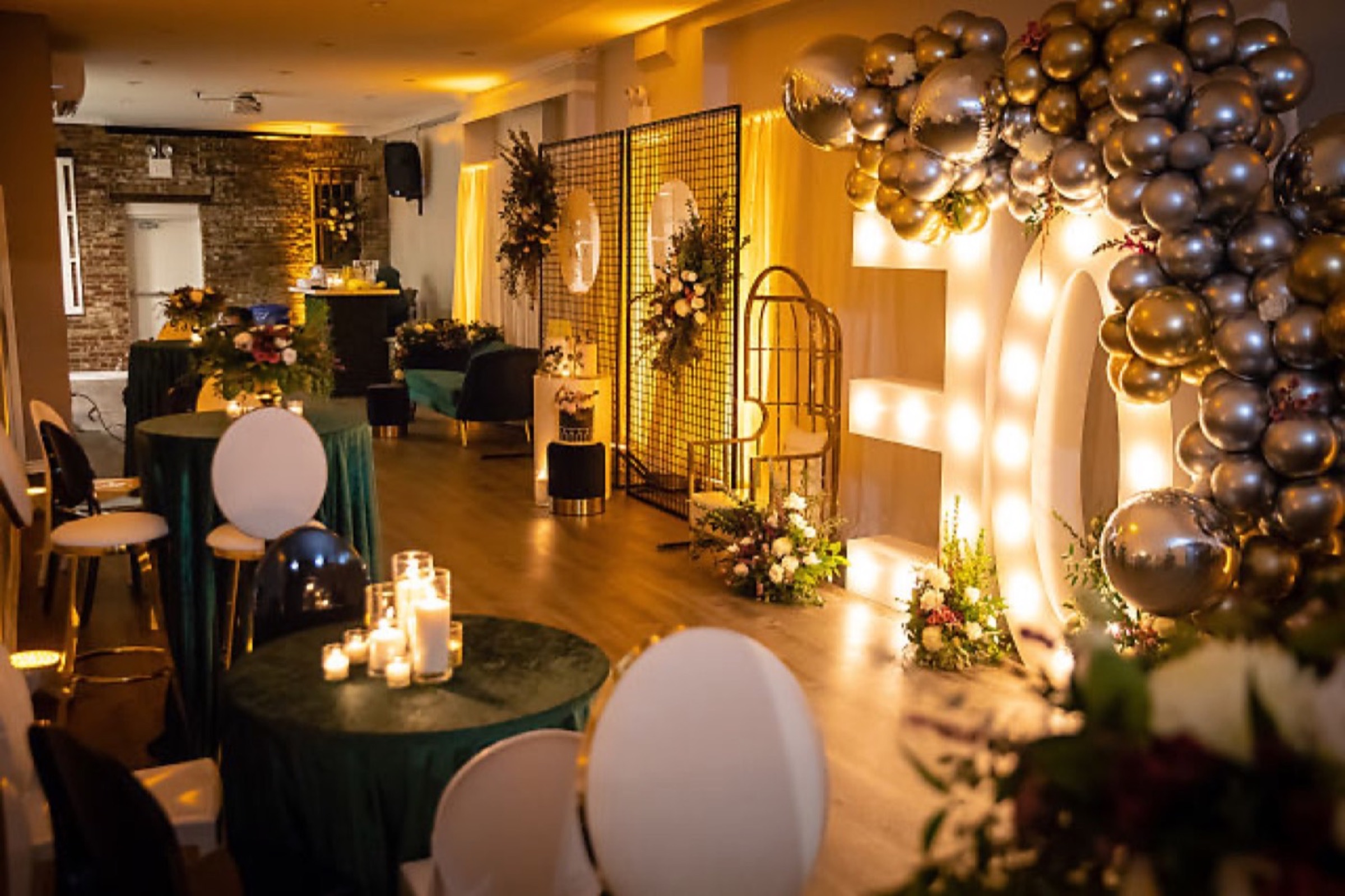 A party room lit in warm gold, with illuminated marquee letters, gold balloons, candlelit tables and floral arrangements.