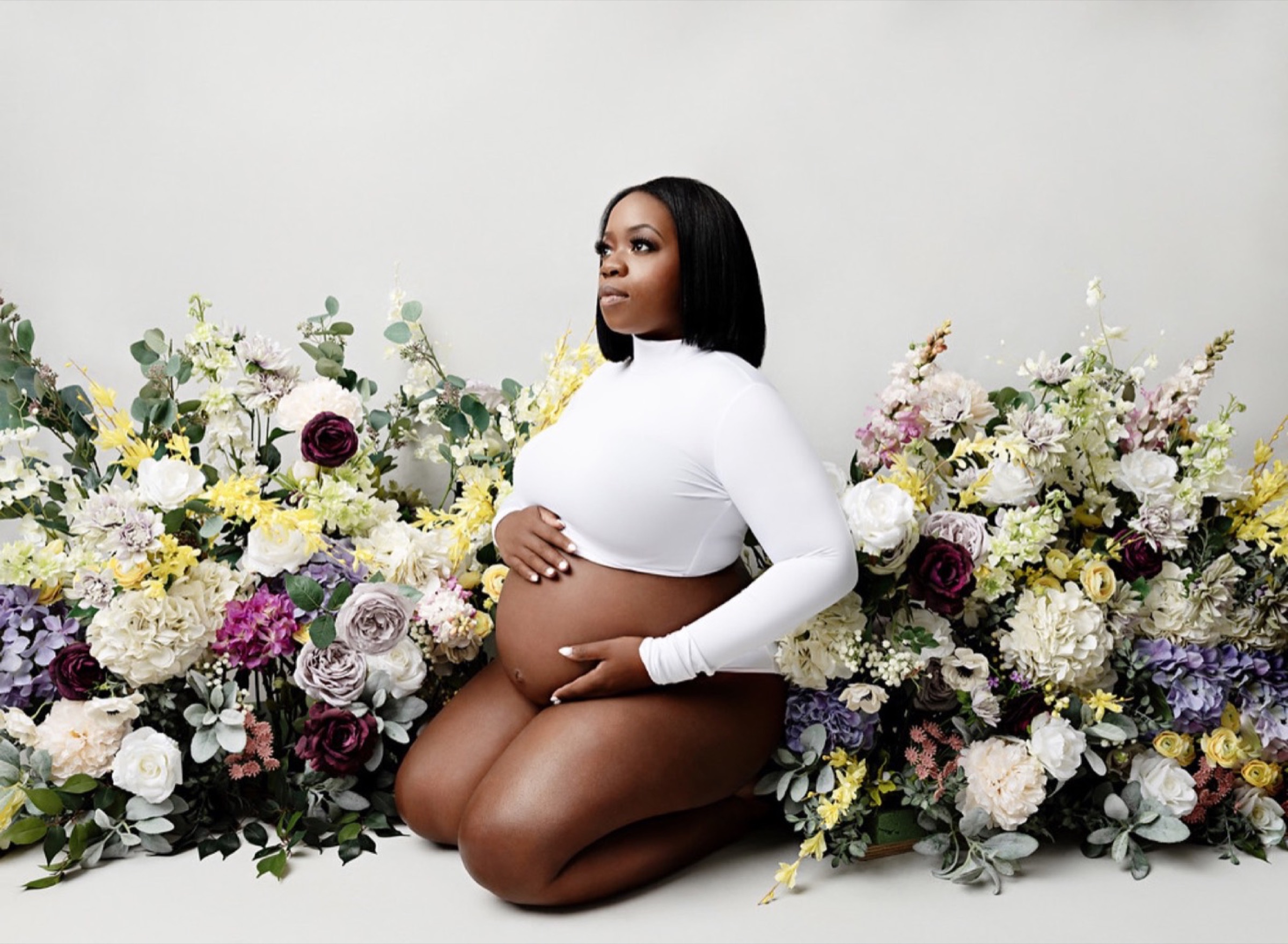 A pregnant woman in a white long-sleeved top kneeling among a wide spread of white, yellow and purple flowers, holding her bump.