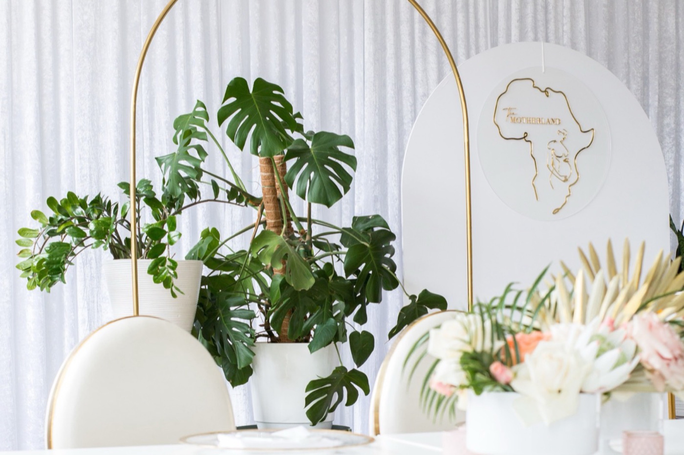 A white event backdrop with gold arches, potted monstera plants and pale floral arrangements.